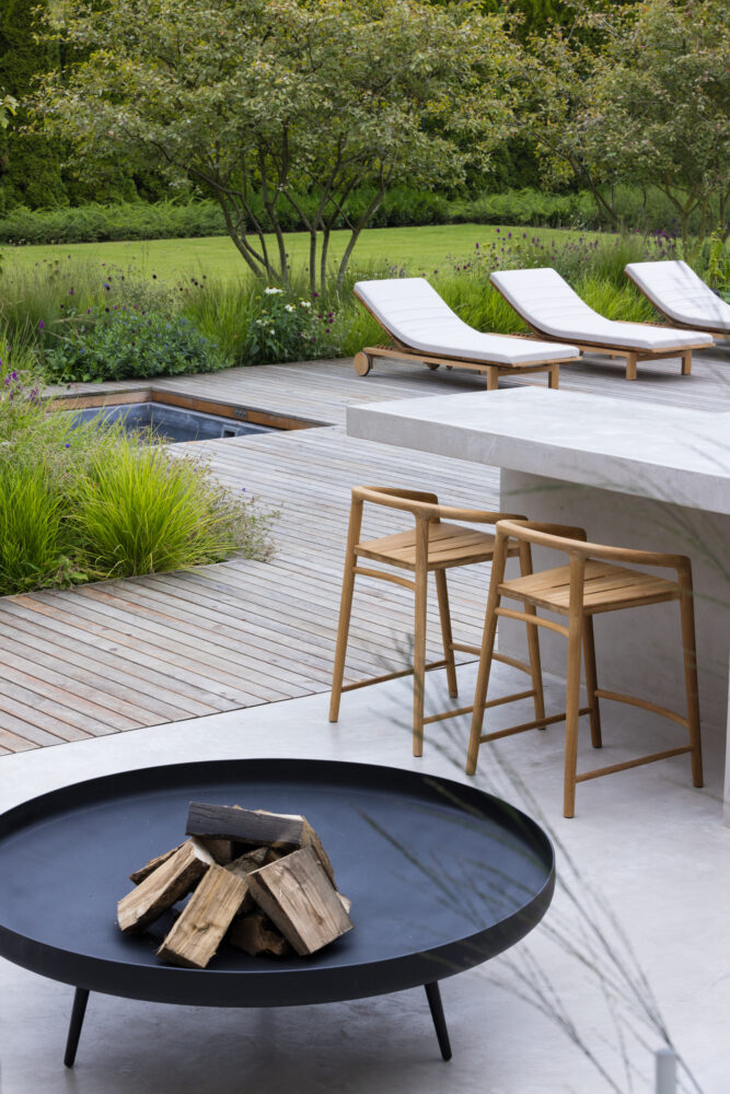 A large Cambridge garden designed and built by Bowles & Wyer, expert garden and landscape designers. The space features a light grey concrete communal bar and grill area with seating, designed to stand out against the pale brick of the house while blending beautifully with the natural green backdrop of trees and planting. mini fireplace stand and wooden decking around the pool